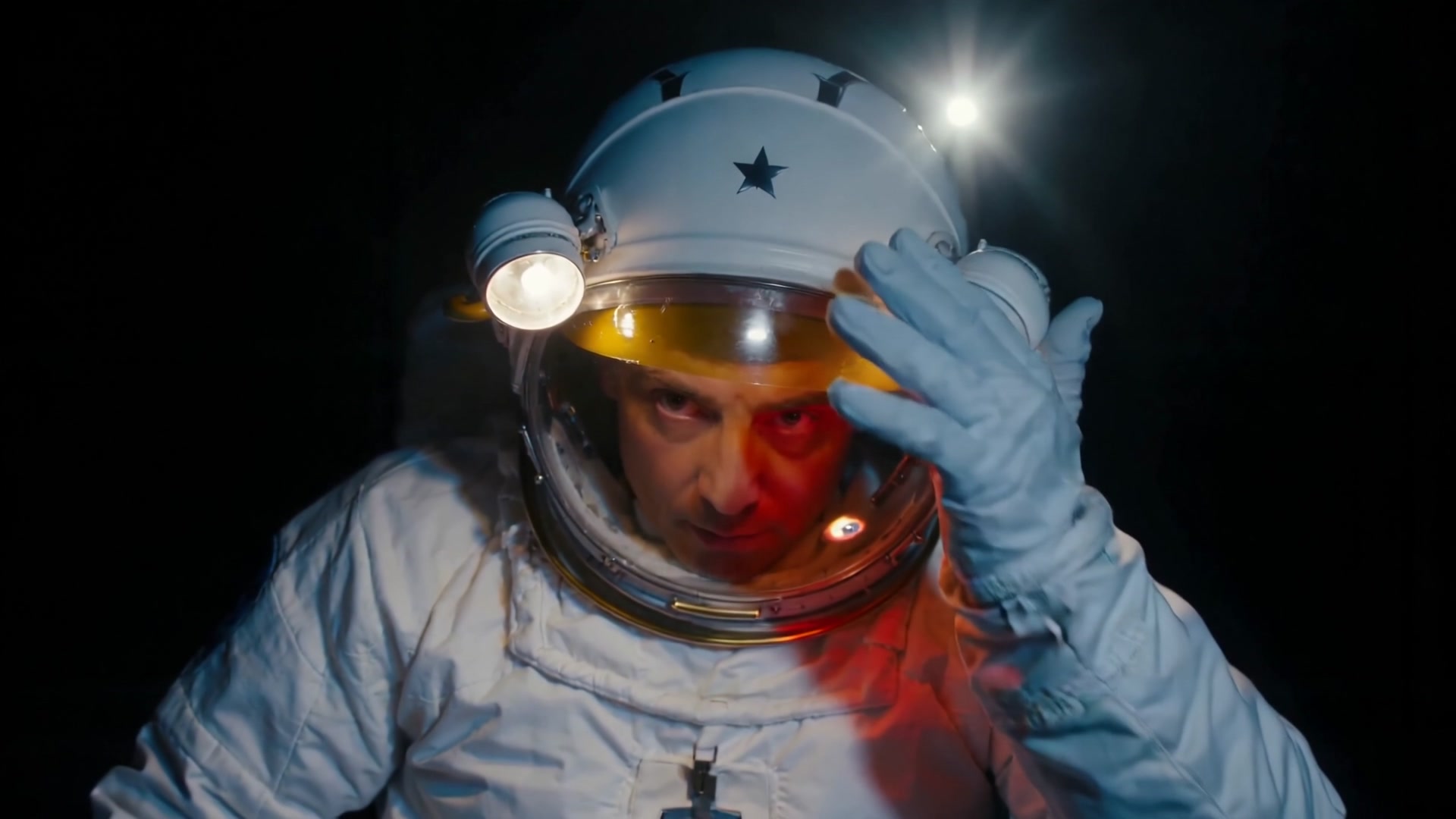 Tether: an astronaut seen through his visor, a gloved hand raised to the helmet, the glass lit red, a single star behind him